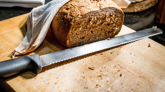 Serrated knife lying on a wooden cutting board next to a partially covered loaf of wholegrain bread, with a rustic kitchen backdrop.